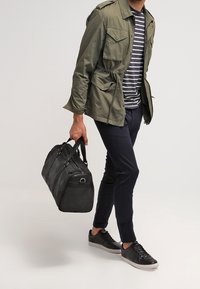 Mężczyzna w zielonej kurtce, w paskowaną koszulę, granatowych spodniach i czarnych sneakersach, idzie, trzymając czarną skórzaną torbę podróżną w prawej ręce.