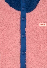 Veste en polaire rose avec des boutons-pression bleus au centre et un petit patch blanc portant l'inscription "TINY" sur le côté droit.