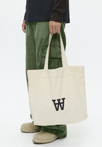 Sac en toile de couleur naturelle avec un grand logo noir en forme de lettre 'W'. Deux poignées robustes ; texture lisse ; exposé avec un pantalon cargo vert et des chaussures beiges.