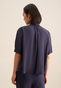 Blusa blu navy a maniche corte con vestibilità ampia, caratterizzata da un collo arricciato e un tessuto fluido. La vista posteriore mostra drappeggi e una texture morbida.