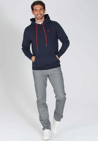 Hoodie marine avec cordons rouges, poche avant et petit logo ; accompagné de pantalons gris et de baskets blanches. Tenue simple et décontractée.