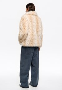 Chaqueta de piel sintética con base beig y patrón de manchas, que presenta un cuello alto y un corte holgado, combinada con pantalones oscuros de pierna ancha.