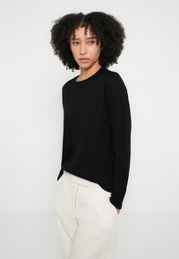 Čierny sveter s dlhým rukávom a okrúhlym výstrihom s hladkou textúrou, v kombinácii so svetlobéžovými nohavicami s textúrou.