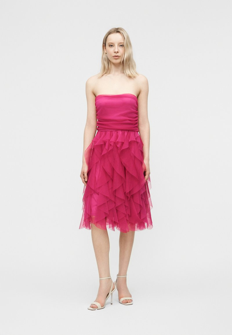 Femme debout portant une robe rose à volants sans bretelles, longueur au genou, et des sandales blanches à talons hauts, sur un fond clair uni.