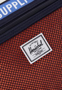 Červený síťovaný batoh s námořnickými akcenty, vybavený zipem a obdélníkovou bílou etiketou, na které je logo 'Herschel Supply Co.'.