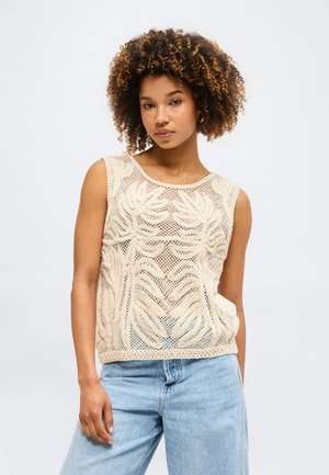 Νεαρή γυναίκα με σγουρά μαλλιά, φοράει μπεζ αμάνικη κορυφή πλέκτη και ανοιχτό μπλε τζιν, στέκεται μπροστά σε ένα λευκό φόντο.