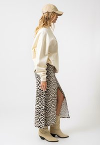 Sweatshirt bej oversized, fustă maxi cu imprimeu leopard și fanta laterală, și cizme din piele de antilopă de stil vestern bej. O șapcă casual completează lookul.