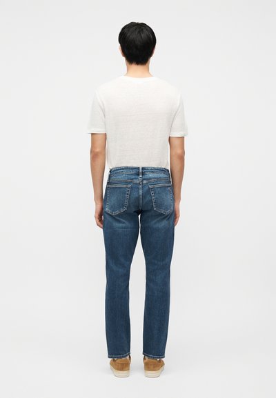 Homme debout de dos, portant une chemise blanche à manches courtes, un jean bleu avec des poches arrière, et des chaussures en daim beige sur fond blanc uni.