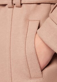 Beżowa tkanina płaszcza o gładkim, teksturowanym wykończeniu. Posiada boczną kieszeń z czystym, szytym brzegiem. Zbliżenie na rękę opartą na kieszeni.