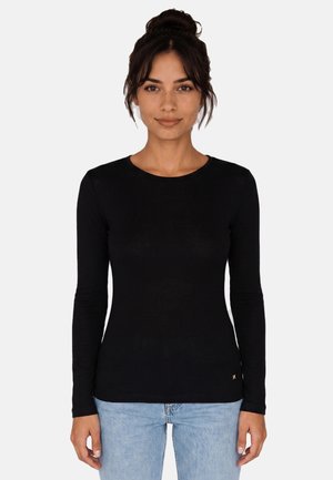 Giovane donna con capelli scuri raccolti in uno chignon, indossa una maglietta nera aderente a maniche lunghe e jeans azzurri, in piedi davanti a uno sfondo semplice.