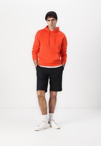 Orange cotton hoodie with a front pocket and silver drawstrings, paired with black shorts, white socks, and white sneakers.