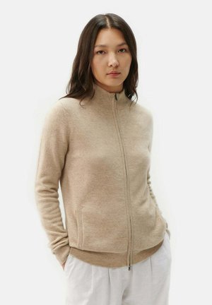 Pull zipé beige avec un col montant, des poignets côtelés et un ourlet côtelé. Fabriqué en matériau doux avec deux poches latérales. Design minimaliste.