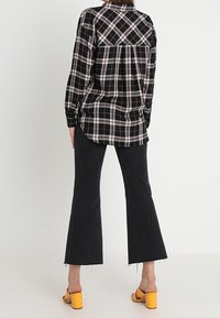 Woman wearing black and white plaid long-sleeve shirt, black cropped flare jeans, and yellow open-toe block heel sandals, standing against plain background.