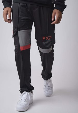 Uomo con pantaloni cargo neri con toppe grigie e rosse, scarpe da ginnastica bianche e felpa nera, in piedi davanti a uno sfondo chiaro uniforme.