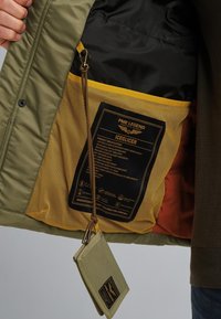 Olivgrön huva jacka med svart foder; har en gul nätficka och en etikett med produktinformation. Nyckelring fäst.