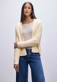 Cárdigan beige y texturizado con diseño de frente abierto, llevado sobre una blusa clara y combinado con jeans de pierna ancha en azul. Look simple y casual.