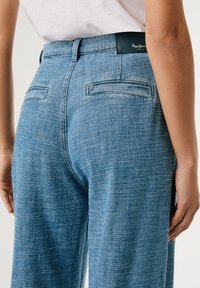 Blue denim trousers with a textured finish, featuring a high waist, side pockets, and a leather brand patch at the back.