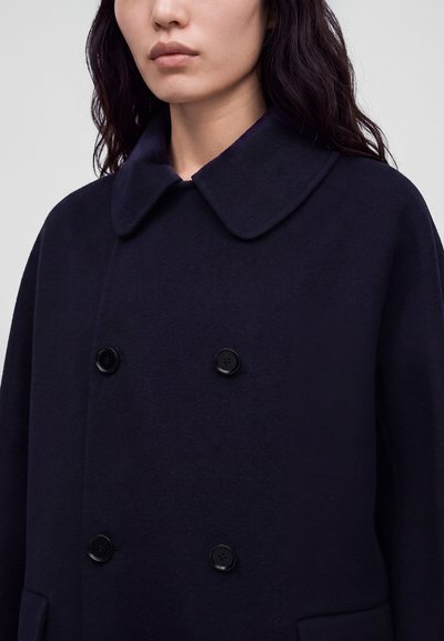 Femme portant un manteau bleu marine foncé à double boutonnage avec quatre boutons et un large col arrondi, visible des épaules jusqu'au milieu du torse.