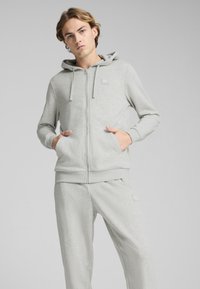 Gaiši pelēks zip-up kapucis ar kengura kabatām un stiepjamu kapuci. Mīksta auduma, ar mazu logotipa uzlīmi uz krūtīm. Redzami saskaņoti sporta bikses.
