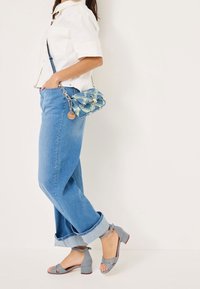 Femme portant un jean bleu retroussé, un t-shirt blanc à manches courtes, des sandales gris à talons blocs, et portant un petit sac bandoulière bleu texturé.