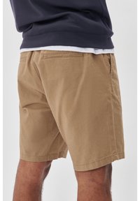 Hnedé bavlnené šortky s voľným strihom, ktoré majú zadné vrecká a obrúbený lem. V kombinácii s tmavomodrým sweatshirtom navrchu.