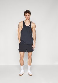 Un débardeur noir à larges bretelles, associé à un short de sport noir. Présente un logo sur la poitrine et des baskets blanches avec des semelles marron.