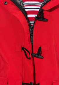 Rode buitenkleding met een rits aan de voorkant, trekkoordaccenten en zwarte toggles; heeft een gestreept binnenhemd. Het materiaal lijkt soepel.