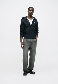 Námornícka modrá mikina na zips s vreckami v štýle klokanky, pod ňou biele tričko, doplnené sivými džínsami s rovnými nohavicami a čiernymi topánkami. Jednoduchý, neformálny outfit.