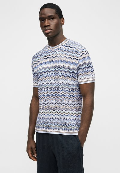 Νεαρός άντρας στέκεται φορώντας ένα κοντομάνικο πουκάμισο με πολύχρωμο σχέδιο ζικ-ζακ και σκούρο υφασμάτινο παντελόνι, με το ένα χέρι στη τσέπη.