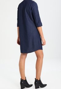 Vestido de mezclilla en azul oscuro con mangas tres cuartos, un corte holgado y un dobladillo redondeado. Se lleva con botas negras hasta el tobillo con detalles recortados.