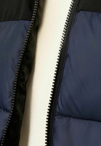 Nedēļas veste ar tumši zilu quilted daļu un melnu audumu plecu daļā, ar redzamu rāvējslēdzēju gar atvērumu.