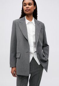 Blazer gri personalizat cu rever mare, închidere cu un singur nasture, două buzunare frontale, purtat peste o cămașă albă cu nasturi. Material texturat.