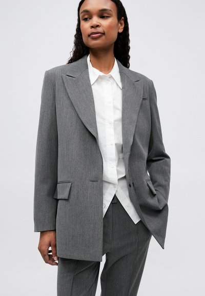 Veste grise ajustée avec grands revers, fermeture à un bouton, deux poches avant, portée sur une chemise blanche à boutons. Tissu texturé.