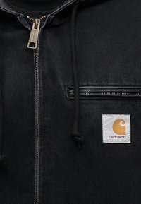Μαύρ ο denim μπουφάν με φερμουάρ μπροστά, κουκούλα με κορδόνι και τσέπη στο στήθος. Περιλαμβάνει μια λευκή ετικέτα με το λογότυπο Carhartt και πορτοκαλί λεπτομέρεια.