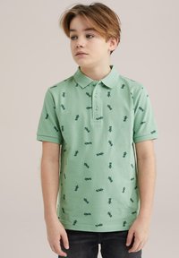 Pikétröja i ljusgrön bomull med korta ärmar; har ett upprepat mönster av mörkgröna motiv. Två-knapps knäppning, klassisk krage.