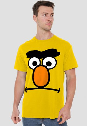 Gelbes Baumwoll-T-Shirt mit einem großen, cartoonhaften Gesicht, schwarzem Augenbrauen, weißen Augen und einer orangen Nase. Rundhalsausschnitt.