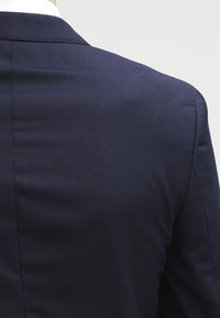 Granatowa marynarka z gładkiego materiału, o zdefiniowanej strukturze i widocznych szwach wzdłuż szwów, z klasycznym kołnierzem.