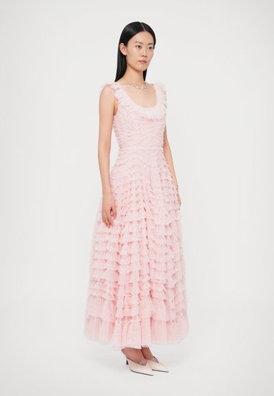 Rózsaszín fodros ruha, testhezálló felsőrésszel, bővülő szoknyával és texturált anyagból. Válltépésekkel és kerek nyakkivágással, fehér magassarkúval párosítva.