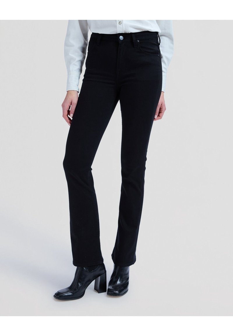 Personne portant un jean noir ajusté à jambes droites, une chemise blanche à boutons et des bottines noires à talons, debout devant un fond uni.