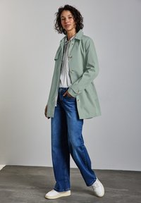 Jasnozielony płaszcz z kołnierzykiem i zapięciem na guziki, w połączeniu z szerokimi niebieskimi dżinsami i białymi trampkami, na neutralnym tle.