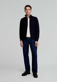 Cardigan in maglia blu navy con colletto alla coreana, chiusura con bottoni, indossato su una camicia color crema, abbinato a jeans blu scuro e scarpe nere. Sfondo neutro.