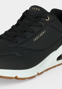 Черни маратонки Skechers с черни връзки, бяла междинна подметка, кафява подметка и логото на марката отстрани и на езика.
