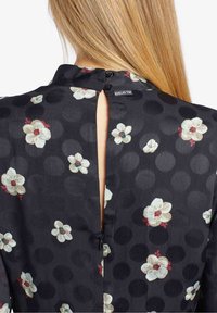 Vue de dos d'une femme aux cheveux blonds portant une robe noire à gros pois foncés et motif floral blanc, avec un col en trou de serrure boutonné.