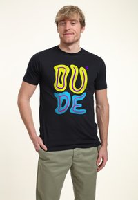 Camiseta negra con el texto "DUDE" en amarillo y azul, de un diseño audaz y colorido, con una textura suave y agradable. Se lleva con pantalones de un color claro.