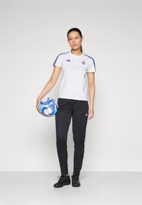 Bílá tričko s krátkým rukávem značky Adidas s modrými pruhy a logem, spárované s černými zúženými kalhotami a černými fotbalovými botami, drží vícebarevný fotbal.
