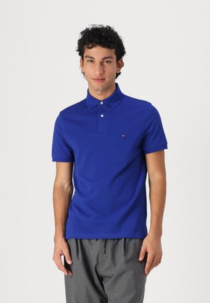 Polo shirt blu royal con colletto, due bottoni e un piccolo logo sul lato sinistro del petto, abbinato a pantaloni grigi.
