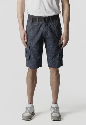 Hombre con pantalones cortos cargo oscuros con estampado y bolsillos laterales, acompañado de un cinturón negro, zapatillas blancas y una camiseta blanca.