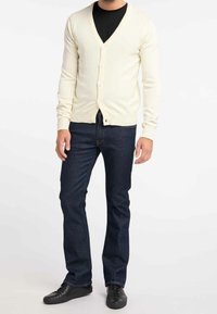 Cremefarbener Cardigan mit V-Ausschnitt, Knopfleiste, gerippten Bündchen und Saum. Trägt darunter ein schwarzes Hemd, kombiniert mit dunkelblauen Jeans und schwarzen Schuhen.