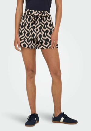 Donna che indossa pantaloncini a vita alta neri e beige con fantasia e cintura annodata, abbinati a sneakers blu navy con strisce bianche.