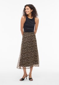 Kvinne med krøllete hår, iført en svart ermeless topp, midi-skirt i dyreprint og svarte hæler, står og smiler med hendene bak ryggen.